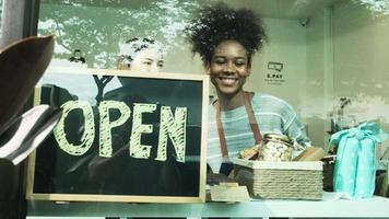 Two young female shopkeepers place open sign board at glass windows display, happy smiles, working at retail store shop, selling consumer products and groceries on shelves, sale occupation lifestyle. video