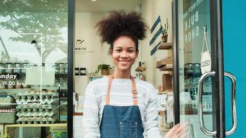 Young African American female shopkeeper opening glass door, happy smile, works at refill store, zero waste, plastic-free grocery shop, eco-friendly SME. Thai language on a label at door means pull.