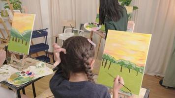 liten flicka koncentrat på akryl Färg bild målning på duk staffli med lärare och barn i konst klassrum, kreativ inlärning med talanger och Kompetens i elementärt skola studio utbildning. video