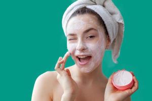 Beauty portrait of young woman in towel on head applying white nourishing mask or creme on face isolated on green background. Skincare cleansing eco organic cosmetic spa relax concept. photo