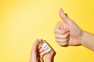 Male hands holding a bottle of covid-19 vaccine and showing a Like gesture on a yellow background. Top view, copy space photo