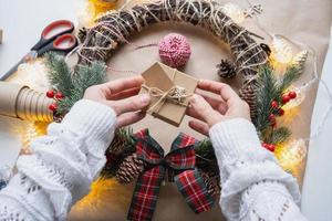 Pack a gift for Christmas and new year in kraft paper, cones, tape, scissors. Tags with mock up, natural decor, hand made, DIY. Festive mood. Flatlay, background photo