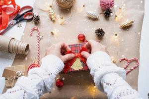 Pack a gift for Christmas and new year in kraft paper, cones, tape, scissors. Tags with mock up, natural decor, hand made, DIY. Festive mood. Flatlay, background photo