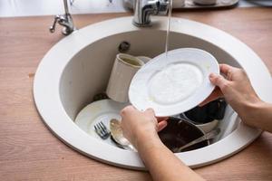 Women's hands wash the plate under the tap water. Dirty dishes in the sink in the kitchen, mess after lunch, too lazy to wash dishes. Kitchen cleaning, detergent, cleaning services photo