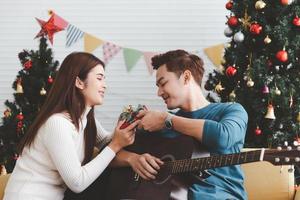 Young adult teenager love couple holding gift present box and looking each other during celebrateing christmas holiday together in living room with christmas tree ornament decoration photo