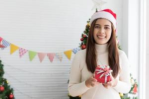 Smiling brunette woman in sweater and Santa hat holding small gift box and rejoices over Christmas decorative white background photo