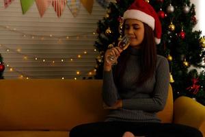 Woman sitting on sofa drinking champagne in living room decorated by lighting and Christmas tree, Holidays and relaxation concept photo