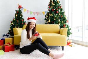 Smiling brunette woman in sweater and Santa hat sitting on the floor in living room holding small gift box and rejoices over Christmas tree decorative background photo