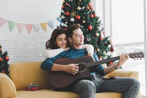 A man playing guitar to woman lover during celebrateing christmas holiday together in living room with christmas tree ornament decoration photo