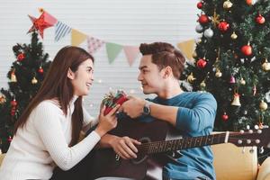 Young adult teenager love couple holding gift present box and looking each other during celebrateing christmas holiday together in living room with christmas tree ornament decoration photo