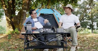 portrait amis d'un homme asiatique heureux assis sur une chaise dans le camp en parlant. ensemble de cuisine cuisine en plein air au sol, voyages, camping, concept de style de vie. video