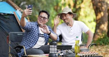 Portrait happy Asian man friends Making a video call with smartphone in camping. Cooking set front ground. Outdoor cooking, traveling, camping, lifestyle concept.