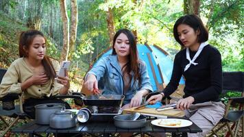 Young asian woman cooking and her friend enjoy to use smartphone take a photo the meal in pan, They are talk and laugh with fun together while camping in nature park video