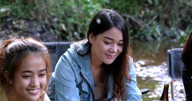 toma de mano, grupo de jóvenes asiáticas sentadas en sus sillas y disfrutando de salpicar agua en el arroyo mientras acampan en el parque natural, hablan y se ríen juntas. video