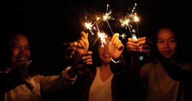 selettivo messa a fuoco mano di asiatico giovane donna e sua amici Tenere fuoco ardente stelle filanti, danza e agitando per giocando con scintillante nel mani, Sorridi insieme su all'aperto nuovo anni festa notte video