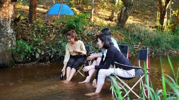 tiro de mano, grupo de jóvenes asiáticas sentadas en sus sillas y disfrutando de salpicar agua en el arroyo mientras acampan en el parque natural, hablan y se ríen juntas. video