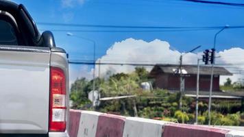 Rear side view of pick up car with turn on brake light. Driving on the road with barrier with red and white color. Background of rural town with tree and lighting pole. Under blue sky and white clouds photo