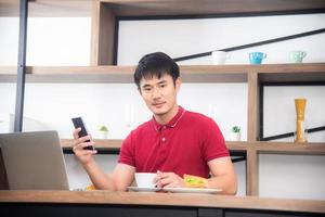 The business man with casual  red t-shirt messaging on mobile phone, drinking coffee and eating sandwich, Young man working on labtop or computer  in the kitchen room, loft style photo