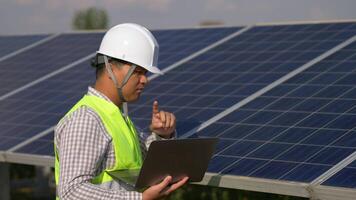 Young Asian technician man walking to checking operation of sun and photovoltaic solar panel and typing on laptop computer while working in solar farm video