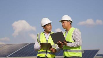 dos técnicos e ingenieros asiáticos usan una computadora portátil mientras verifican la eficiencia del sol para examinar la construcción de paneles solares y hablar juntos video