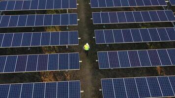 Slow motion shot, Aerial drone view, Flight over solar panel farm and young engineer wearing white helmet talking on smartphone and walking between solar panels row video