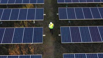 Slow motion shot, Aerial drone view, Flight over solar panel farm and young engineer wearing white helmet talking on smartphone and walking between solar panels row