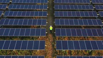 Slow motion shot, Aerial drone view, Flight over solar panel farm, back of young engineer talking on smartphone and walking between solar panels row video