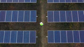 Aerial drone view, Flight over solar panel farm and young engineer wearing white helmet talking on smartphone and walking between solar panels row video