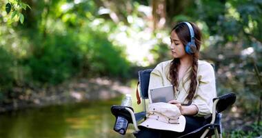 jonge aziatische vrouw zit in een stoel in de buurt van de beek en luistert graag naar muziek van een tablet met een draadloze koptelefoon tijdens het kamperen in het bos video