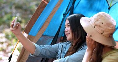 Handheld shot, Close up, Young Asian pretty woman and her girlfriend sitting at front of tent, use mobile phone take photo and checking during camping in forest with happiness together video