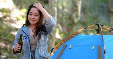 Young beautiful Asian woman relaxing while camping in forest, standing to dancing and drinking beer with happy video