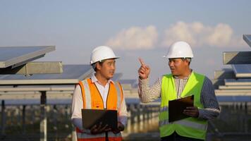 Two Asian young engineers walking along rows of photovoltaic panels in solar farm, They use laptop computer and talking together video