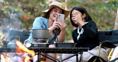 Handaufnahme, Vorderansicht Junge asiatische hübsche Frau und ihre Freundin sitzen vor dem Zelt und überprüfen das Foto im Handy während des Campings im Wald mit Glück zusammen video