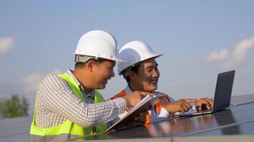 asiatischer inspektor ingenieur mann hält kontrollbrett und junger technologiemann benutzt laptop-computer, zwei ingenieure diskutieren während der arbeit im solarpark, solarpanelstation im hintergrund video