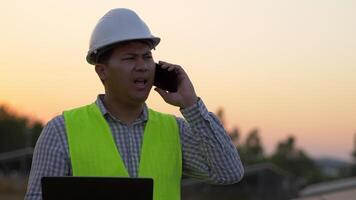 Young Asian Technician Engineer man hold laptop computer and upset during talking on smartphone while check efficiency of sun for examination on solar panel construction on sunset video