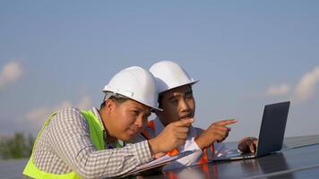 asiatischer inspektor ingenieurmann und junger technologiemann verwenden laptop-computer, zwei ingenieure diskutieren während der arbeit im solarpark, solarpanelstation im hintergrund video