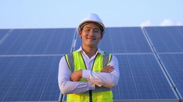 porträt asiatischer junger ingenieur trägt einen weißen helm, der mit einem lächeln die arme verschränkt und auf die kamera des solarparks schaut, solarpanel im hintergrund video