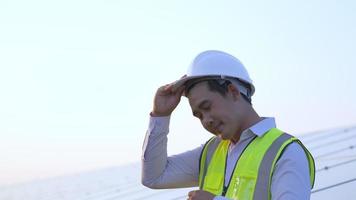 Der junge Techniker-Ingenieur nimmt den weißen Helm ab und wischt sich in den Pausen heiß und müde den Schweiß im Gesicht ab. Sonnenkollektoren im Hintergrund video
