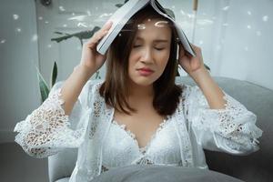 mujer asiática usa camisón blanco que sufre tensión ocular leyendo un libro sentado en un sofá en la noche. foto