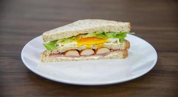 Sandwich on white plate and wooden table, made from bread, sausage, fried egg and vetgetable is American breakfast or fast food is unhealthy food or junk food photo