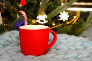 taza roja en blanco con árbol de navidad en el fondo, taza de té o café con decoración de navidad y año nuevo, maqueta horizontal con taza de cerámica para bebidas calientes, plantilla de impresión de regalo vacía. foto