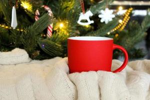 taza roja en blanco con árbol de navidad en el fondo, taza de té o café con decoración de navidad y año nuevo, maqueta horizontal con taza de cerámica para bebidas calientes, plantilla de impresión de regalo vacía. foto