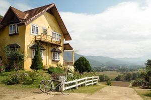 pintoresco paisaje rural con casa amarilla con jardín y bicicleta en primer plano y montañas y campos en segundo plano. foto