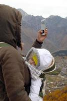 retrato de una joven madre con su pequeña niña dormida en un portabebés sobre un fondo de montañas otoñales. la mujer está tomando una foto desde el teléfono inteligente.