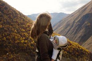 retrato de una joven hermosa mujer con su pequeña hija dormida en un portabebés ergo sobre un fondo de montañas otoñales. montañas del cáucaso, georgia. foto
