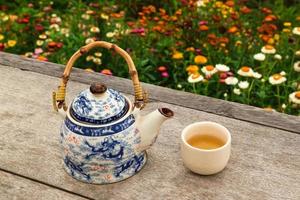 Chinese teapot and cup with green tea on a wooden table on a background of bright colorful flowers, top view. Pai, Thailand. photo