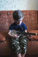 lindo niño europeo, de 5 años, toca una guitarra eléctrica de juguete en casa en el sofá foto
