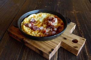 Fried eggs and bacon in a frying pan on a wooden stand on a rough wooden table, breakfast in a restaurant, side view photo