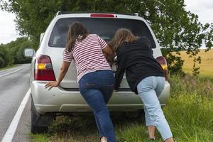 un coche roto. dos mujeres jóvenes empujan un camión roto en la carretera, una avería, contra el fondo de un campo amarillo foto