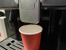 Red cardboard coffee cup into which delicious invigorating hot black Americano coffee is poured from the coffee machine in the office in the morning photo